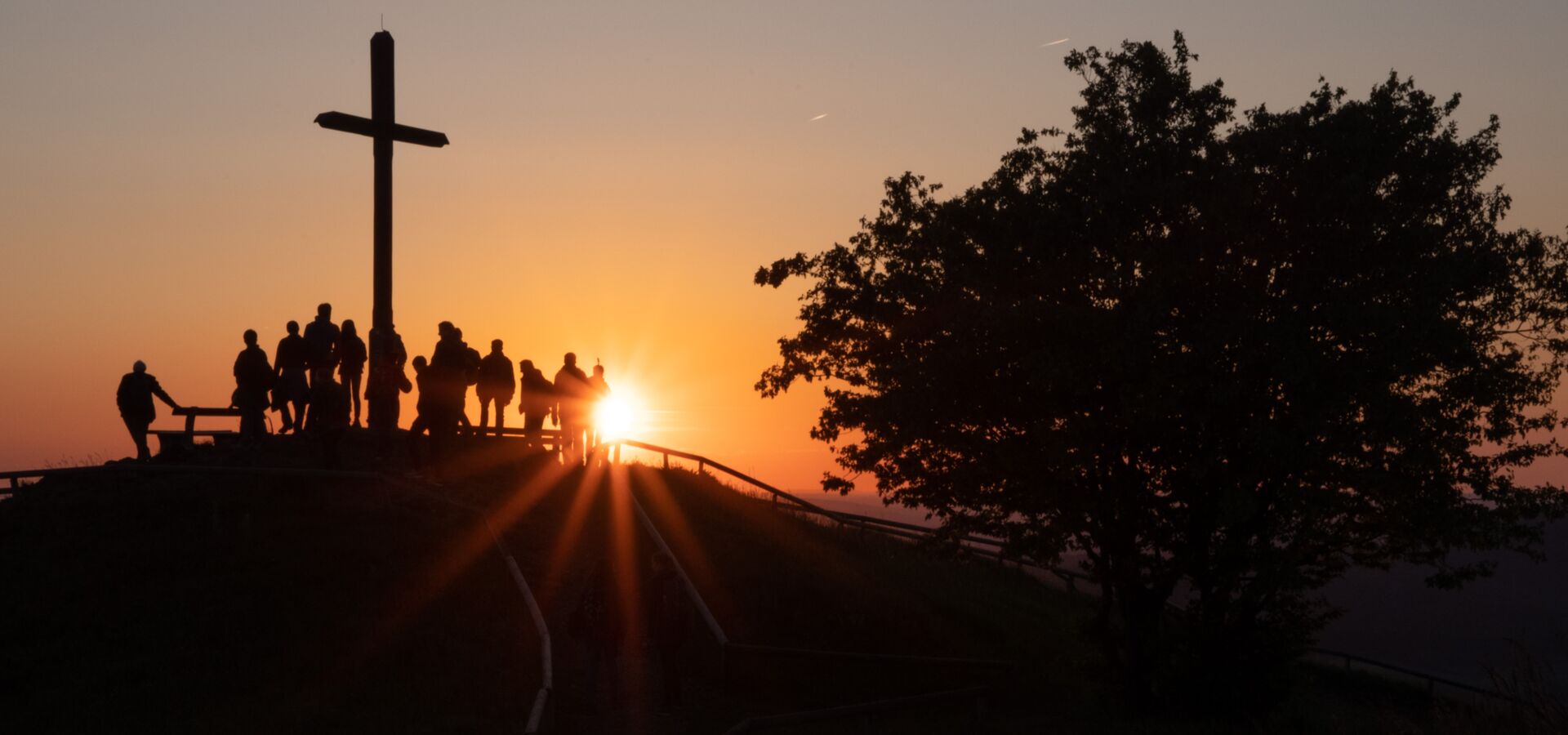 an einem Gipfelkreuz steht eine Gruppe von Leuten; die Sonne geht unter